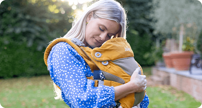 Maman câlinant son enfant installé dans un porte-bébé Joie Savvy, coloris jaune moutarde